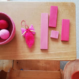 Vier rosa und pinke Holzbausteine von Glückskäfer liegen neben einer kleinen Fee aus Filz, und einem Korb gefüllt mit zwei Häkelbällen von Kokomoo, ebenfalls alles pink.