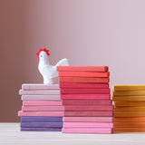 Die bunten Holzbausteine von Glückskäfer sind in drei Stapeln nebeneinander sortiert aufgestellt, Lila- und Rosatöne ganz link, in der Mitte Rosa- und Rottöne und ganz rechts Gelb und Orange. Auf dem linken Stapel sitzt eine kleine Henne aus Filz.