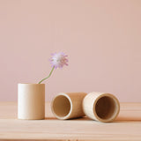 Drei glatte Holzbecher Cups von Grapat, einer steht, mit einer Blume darin, zwei liegen daneben.