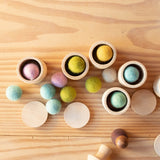 Glatte Holzdosen oder Holzbecher, gefüllt mit pastellfarbigen kleinen Filzkugeln, das freie Spiel im Pikler Spielraum.