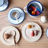 Aufgetischt aus der Puppenküche: Emaille Geschirr, Holzteller, Kochlöffel und Teller aus Holz, ein Holzei im Eicherbecher. Kleine Spielfiguren liegen auch in Topf und Teller.