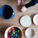 Kinderhände, ein blaues Körbchen, ein Korb gefüllt mit bunten Mandala Steinen aus Holz und daneben Holzteller und Schüsseln aus Holz, alles bereit für das freie Spiel.