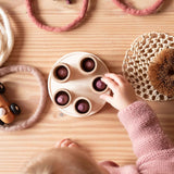 Ein Kleinkind spielt mit Mandala Steinen in gerillten Bechern on Grapat auf einem Holzteller. Daneben liegen Filzringe.