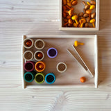 Zwei Holztabletts, in dem oben sind orange Mandala Steine aus Holz in Kürbisform, im größeren Tablett darunter sind Becher, eine Mini-Holzzange zum Greifen, und Holzbecher, teilweise schon befüllt mit den Mandala Kürbissen.