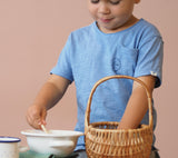 Ein Kind spielt kochen, mit der rechten Hand rührt es in einer kleinen Emailleschüssel, mit der rechten Hand greift es in den kleinen Korb von Kokomoo.