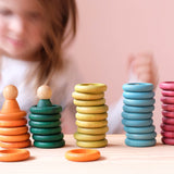 Die Holzplättchen und Holzringe des Grapat "Nins, Rings & Coins" Holzfiguren Spielset nebeneinander gestapelt aufgestellt, dahinter ein Kind.