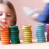 Die Holzplättchen und Holzringe des Grapat "Nins, Rings & Coins" Holzfiguren Spielset nebeneinander gestapelt aufgestellt, ein Kind setzt einen orangen Holzring auf den Stapel.