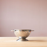 Edelstahl-Seiher von Glueckskaefer mit vielen Loechern steht auf Holzplatte