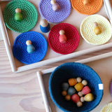 Sechs runde, unifarbene Häkeldeckchen liegen nebeneinander auf einem Holztablett. Auf jedem Deckerl liegt eine kleine Holzfigur von Grapat, die farblich zum Deckchen passt.