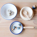 Draufblick auf eine weiße, kleine Emailleschale mit blauem Rand. Darin liegen Steine. Daneben steht ein Holzteller, darauf eine Naturholzschale mit Kochlöffel. Davor steht eine Emaillepfanne mit 3 kleinen, blauen Holzfiguren - BabyNins von Grapt, die darin liegen.