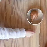 man sieht von oben auf eine Holzfläche. Der Arm eines Kindes streckt sich nach einer runden, flachen Holzschale in der die Rassel Kugelrund liegt.