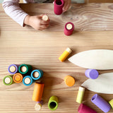 Grapat Lolas in Erdönen und Becher liegen oder stehen kunterbunt durcheinander auf dem Boden. Kinderhände halten einen himbeerfarbenen Becher und eine farblich passende Spielfigur.