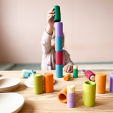 Ein Kind baut einen hohen bunten Turm aus den zylinderförmigen Bechern von Lola in Erdtönen. Davor stehen und liegen Lolas und anderes Spielmaterial auf dem Tisch.