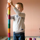 Ein Kind baut einen sehr hohen Turm aus den zylinderförmigen Bechern von Lola in verschiedenen Erdtönen, der schon fast größer ist, als das Kind selbst im Stehen.