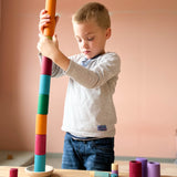 Ein Kind hält einen sehr hohen Turm aus den zylinderförmigen Bechern von Lola in verschiedenen Erdtönen, der Turm ist in eine kleine Holzschale gebaut.