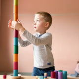 Ein Kind hält schützend seine Hände um einen sehr hohen Turm aus den zylinderförmigen Bechern von Lola in verschiedenen Erdtönen, hoffend, dass er nicht zusammen stürzt.