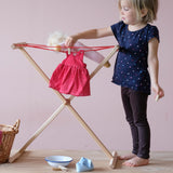 Das rote Tupfenkleid der Stoffpuppe Lotti hängt auf der kleinen Wäscheleine für Kinder, die Puppe liegt daneben, ein Kind löst grad die Wäscheklammern vom Kleid.