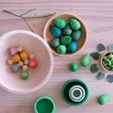 Sechs kleine Holzfiguren zum Spielen in einer Holzschale, in der kleineren Schale daneben liegen Mandala Bäume von Grapat.