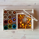 Ein Bick von oben auf ein Natur-Holztablett. Darauf sieht man eine quadratische, flache Holzbox, in der sich orange-gelbe kleine Holzkürbisse von Grapat befinden. Eine Mini-Holzzange liegt darauf. Daneben stehen 12 kleine Holzbecher in natur und in Erdtönen, die teilweise mit den Kürbissen gefüllt sind.