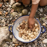 Auf einem Kiesstrand steht eine Emailleschüssel gefüllt mit Mandalasteinen von Grapat aus naturbelassenem Holz.
