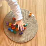 Eine Kinderhand greift nach den Mandala Pilzen von Grapat "Mushrooms Rainbow", die auf einem runden Tischset liegen.