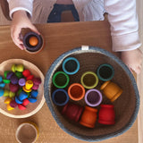 Ein Holzschale mit Mandalasteinen Pilze von Grapat namens "Mushrooms Rainbow", daneben ein Körbchen mit Grapat Mates Holztöpfen. Ein Kind spielt damit.