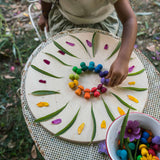 Auf der runden Holzplatte natur von Grapat legt ein Kind ein Mandala mit echten Blüten, Blättern und den Mandala Pilzen Regenbogen von Grapat namens"Mushrooms Rainbow".