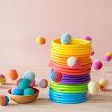 24er-Set Motorikringe in sechs Regenbogenfarben aufeinander gestapelt, drumherum fliegen die süßen Häkelbälle von Kokomoo in verschiedenen Farben.