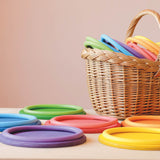 Die Motorikringe in den sechs Regenbogenfarben liegen auf sechs flachen Holztellern, die farblich jeweils genau passen, dahinter steht ein Korb mit noch mehr Motorikringen.