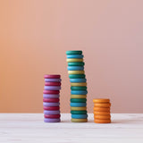 Drei Stapel Coins Holzplättchen in Erdtönen: ganz links 12 in Lila-Violetttönen, in der Mitte Grün- und Blautöne und ganz rechts ein Stapel mit 6 orangen Plättchen.