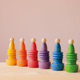 Sechs Grapat Nins Spielfiguren in 6 Regenbogenfarben stehen auf 6 Coins Holzplättchen-Stapel, die wiederum auf Bechern und Schalen von Grapat stehen.