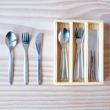 Ein Löffel, eine Gabel und ein Messer für Kinder liegen neben einem Holzkasten, in dem in den drei Fächern weitere 3 Löffeln ganz links, 3 Gabeln in der Mitte und 3 Messer rechts liegen.