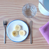Kleine Jause für kleine Kinder: drei Scheiben Banane auf einem kleinen Emailleteller, links und rechts Gabel und Messer, ein Pikler Trinklernglas davor.