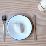 Frühstück in der Puupenküche: zwei Steine auf einem kleinen weißen Teller mit blauem Rand, links und rechts Gabel und Messer, ein Holzei im Eierbecher.