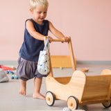 Ein Kind schiebt den Puppenwagen aus Holz von Lammetal, über den Griff hängt rechts eine kleine Stofftasche.
