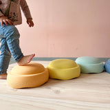 Am Boden liegen Stapelsteine® Original in den pastelligen Farben orange, gelb und mint. Ein Kind steigt mit dem Fuß auf den ersten Stapelstein.