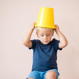 Ein Kind setzt sich einen gelben Sandspielkübel ohne Henkel auf den Kopf.