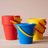 Fünf rote gestapelte Sandeimer, ein blauer Sandkübel und drei gelbe, ebenfalls gestapelt stehen nebeneinander.