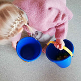 Ein Kind trägt in jeder Hand einen blauen Sandspielkübel mit gelben Henkel.