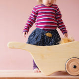 Ein kleines Kind steht vor derHolz Schubkarre von Lautenbach und schaut auf die Puppe, die im Holzwagen liegt.