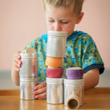 Ein Kind stapelt zwei Schraubdosen von Kokomoo, im oberen sind Grapat Holzplättchen. Daneben ein fertiger Teil des Bauwerks aus Dosen, Grapat Schälchen und Plättchen.