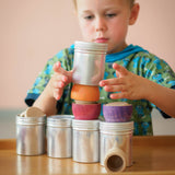 Ein Kind löst seine Hände vorsichtig von der Blechdose mit Schraubverschluss, die er soeben auf ein Bauwerk aus Dosen, Grapat Schälchen und Plättchen gestapelt hat.