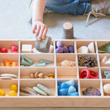 Der Tinker Tray Setzkasten von Grapat aus Holz liegt am Boden, gefüllt mit allerlei Mandalas, Holzfiguren, Steinen, Zapfen, Blättern und anderen Schätzen. Eine Kinderhand möchte gerade einen weiteren Stein einsortieren.