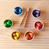 Grapat "Bowls and Marbles" Sortierspiel von oben mit den sechs Schälchen in Regenbogenfarben, befüllt mit den 36 kleinen Holzkugeln und in der Mitte liegt die kleine Holzzange.