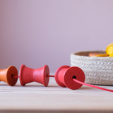 Einige Grapat "Spools" Spulen aus Holz in 6 bunten Regenbogenfarben sind auf einer dicken, roten Schnur aufgefädelt.