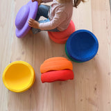 Ein Kind sitzt auf einem rosa Stapelstein und hat ein lila Balanceboard in der Hand, daneben verschieden Stapelsteine in gelb, orange, rot, blau und grün.