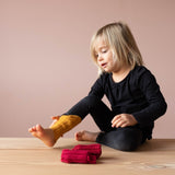 Ein Kind bestaunt die gelbe Stulpe auf seinem rechten Knöchel und der Wade, vor ihm liegt ein Paar rote Stulpen.