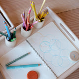 Auf einem großen Holztablett von Kokomoo stehen drei Holzbecher mit Farbstiften und liegt ein offener Block. Es wurden Kreise mit blau und orange gemalt, rundum Holzplättchen.