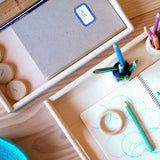 Zwei Holztabletts von oben sind beide teilweise zu sehen, eines größer mit Farbstiften und offenem Block, ein kleineres oberhalb mit einem geschlossenen A5 Heft und Holzplättchen mit Zahlen.