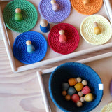 Zwei Holztabletts von oben sind beide teilweise zu sehen, eines größer mit sechs bunten gehäkelten Untersetzern mit farblich passenden Grapat Nins darauf, ein kleineres unterhalb mit einem blauen Körbchen mit weiteren Grapat Holzfiguren darin.